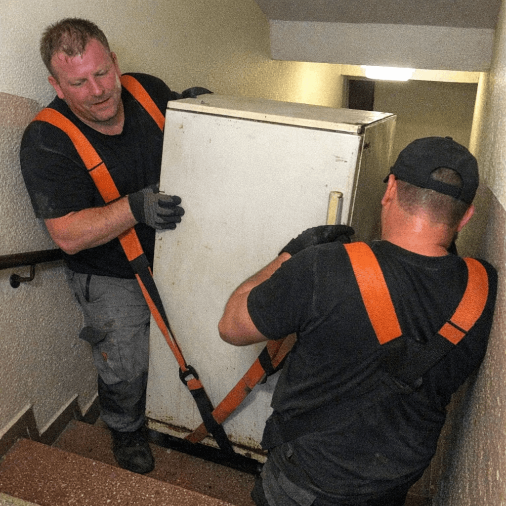 Zwei Mitarbeiter tragen eine alte Gefriertruhe mit Gurten eine enge Kellertreppe hinauf
