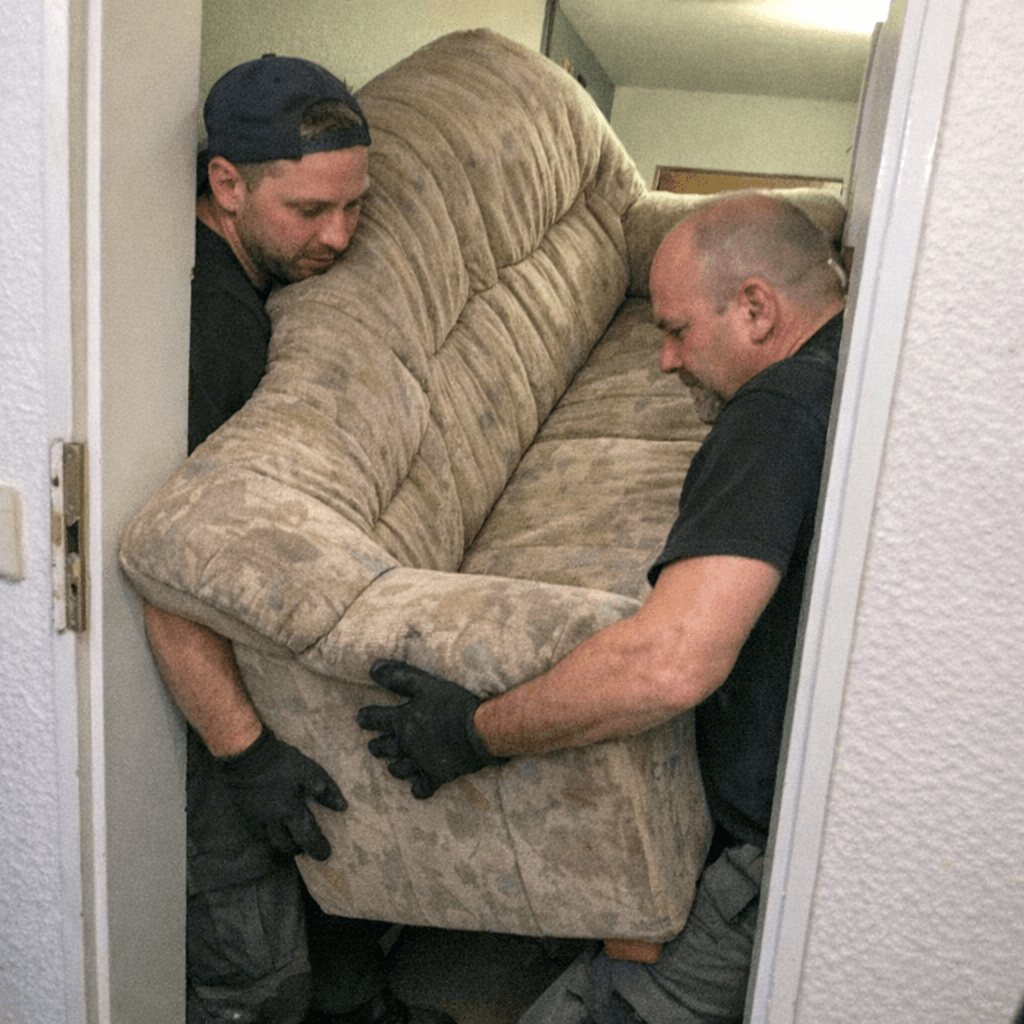 Zwei Mitarbeiter manövrieren ein altes Sofa vorsichtig durch eine enge Haustür
